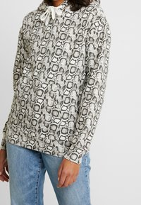 Mikina s kapucí v hadím vzoru, v šedých a bílých barvách, s přední kapsou a nastavitelnými šňůrkami na límci.