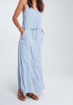 Kobieta ubrana w bezrękawową, jasnoniebieską maxi sukienkę na guziki z paskiem w talii i brązowe plecione płaskie sandały, stojąca z rękami w kieszeniach.