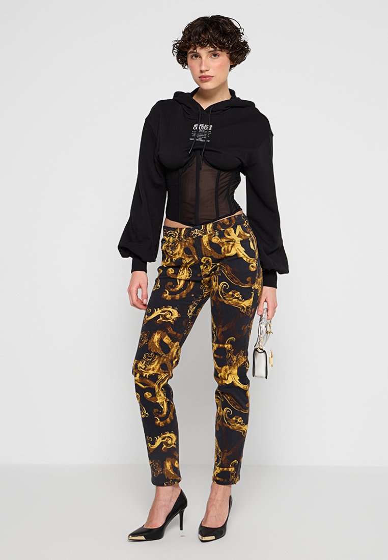 Sudadera recortada negra con cordón, pantalones negros y dorados con estampado, y tacones altos negros. El conjunto presenta un detalle de corsé transparente.