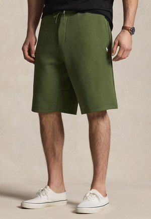 Uomo che indossa pantaloncini verde oliva fino al ginocchio, maglietta nera, sneakers bianche e orologio in pelle marrone, in piedi su uno sfondo beige.
