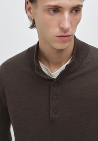 Maglione marrone a maglia con una texture a coste, caratterizzato da un colletto a scialle e tre bottoni neri sul davanti. Indossato sopra una camicia chiara.