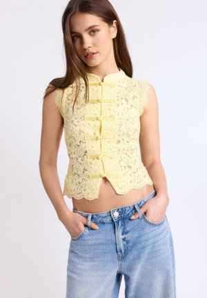 Jeune femme portant un chemisier en dentelle jaune sans manches avec des fermetures par nœuds et un jean bleu clair taille haute, les mains dans les poches, fond neutre.