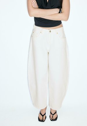 Kobieta ubrana w luźne białe spodnie, czarny bezrękawnik, czarna bransoletka i czarne sandały na obcasie, stojąca z założonymi ramionami.