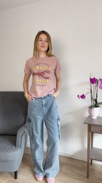 T-shirt grafica rosa con stampa di aragosta e testo. Indossato con pantaloni cargo in corduroy blu chiaro. Texture morbida e vestibilità rilassata.