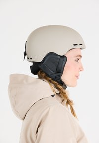 Beige Helm mit glatter, matter Oberfläche, schwarzen gepolsterten Ohrabdeckungen und Belüftungsschlitzen. Getragen mit einer beige Kapuzenjacke. Seitenansicht.