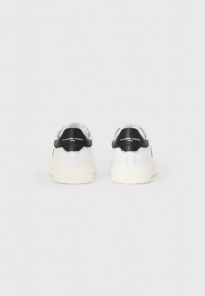 Weiße Sneaker mit glatter Oberfläche, schwarzen Fersenelementen, gepolsterter Knöchelklette und strukturiertem weißen Gummisohle; Logo auf der Ferse zu sehen.