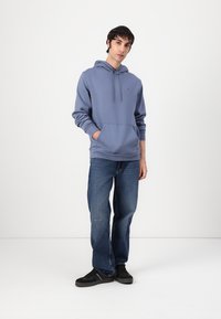Felpa di cotone blu con cappuccio regolabile, tasca a marsupio e polsini a coste, abbinata a jeans blu scuro e sneaker nere.