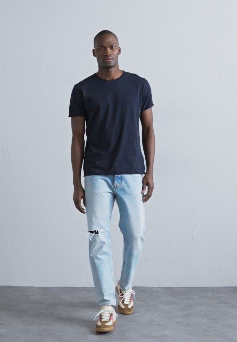 T-shirt en coton noir, jean en denim délavé bleu clair et baskets marron avec accents rouges. Le modèle se tient devant un fond neutre.