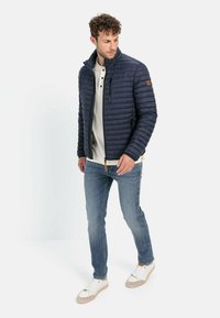Marineblå quiltet jakke med høj krave, lynlåslommer og kontrasterende tan detaljer, kendt med lyseblå jeans og hvide sneakers.