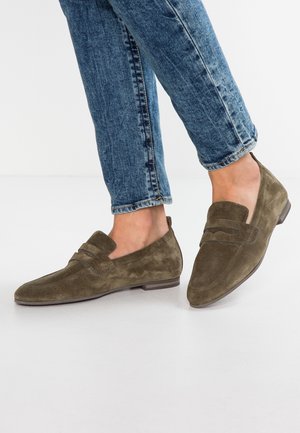 Person trägt blaue Jeans und olivgrüne Wildleder-Loafer, steht auf einer weißen Fläche mit einem leicht gehobenen Fuß.