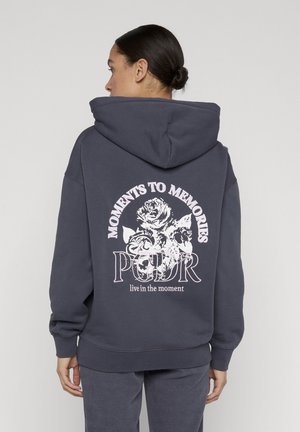Donna con felpa grigia scuro con cappuccio alzato, vista posteriore che mostra un disegno floreale bianco e il testo "MOMENTS TO MEMORIES, PGDR, vivi il momento."