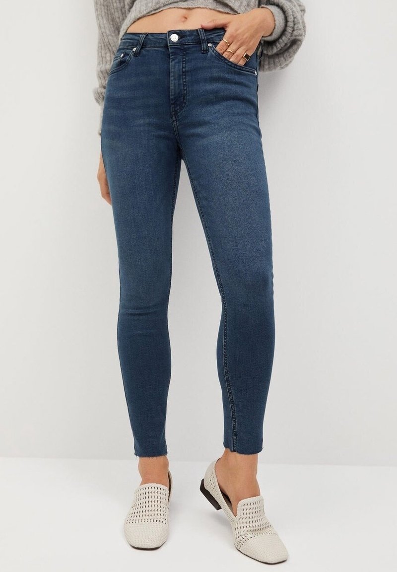 Femme portant un jean skinny bleu foncé, un pull court gris et des chaussures blanches tissées sans lacets, debout devant un fond clair uni.