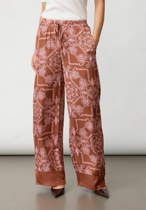 Kobieta nosząca szerokie, rdzawo-brązowe spodnie w różowe wzory paisley, zestawione z brązowymi, spiczastymi butami oraz białą bluzką.