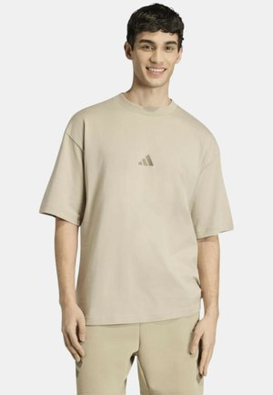 Giovane uomo sorridente, indossa una T-shirt Adidas beige chiaro e pantaloncini coordinati su uno sfondo chiaro uniforme.