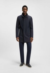 Cappotto impermeabile blu scuro con colletto alto, leggera lucentezza e tasche laterali. Abbinato a pantaloni coordinati e scarpe slip-on con suole beige.