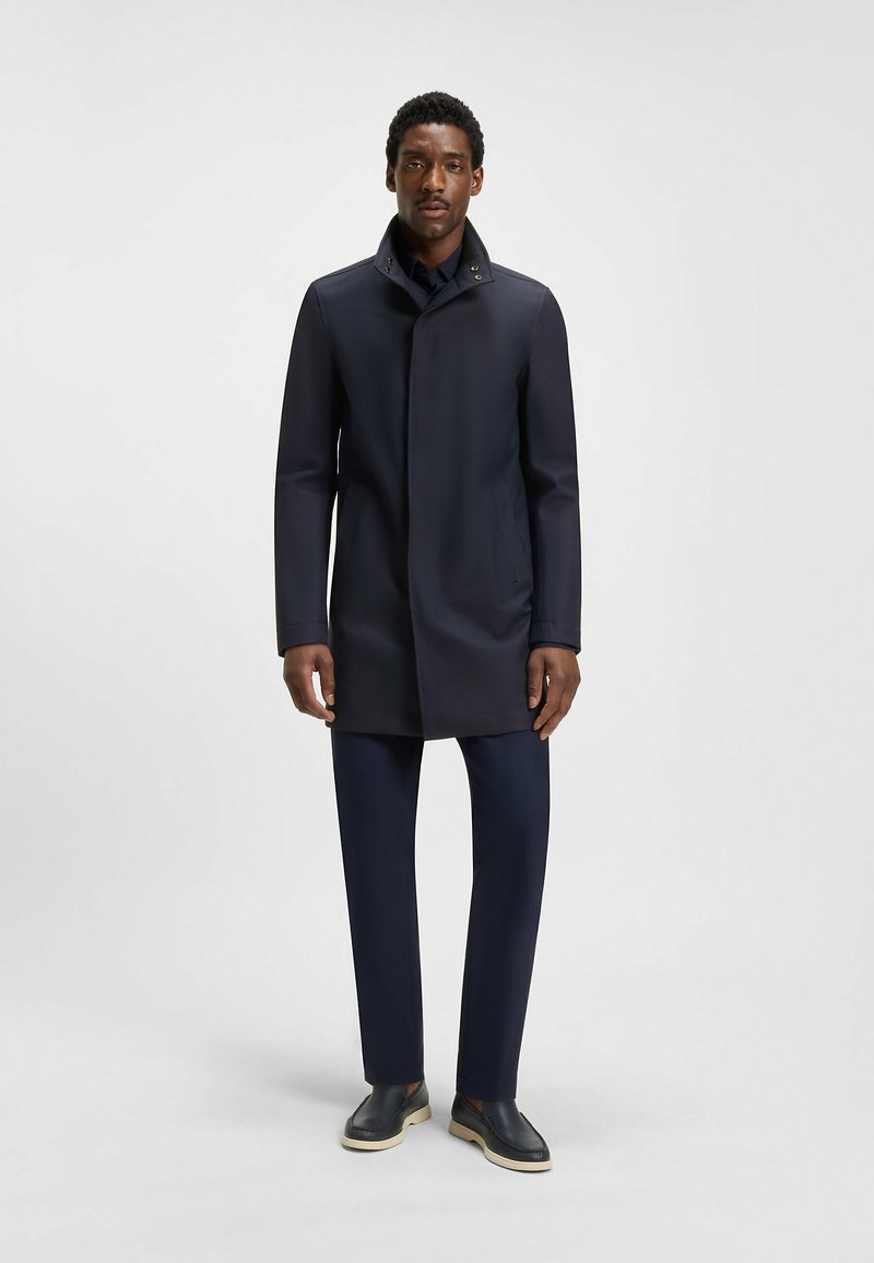 Cappotto impermeabile blu scuro con colletto alto, leggera lucentezza e tasche laterali. Abbinato a pantaloni coordinati e scarpe slip-on con suole beige.