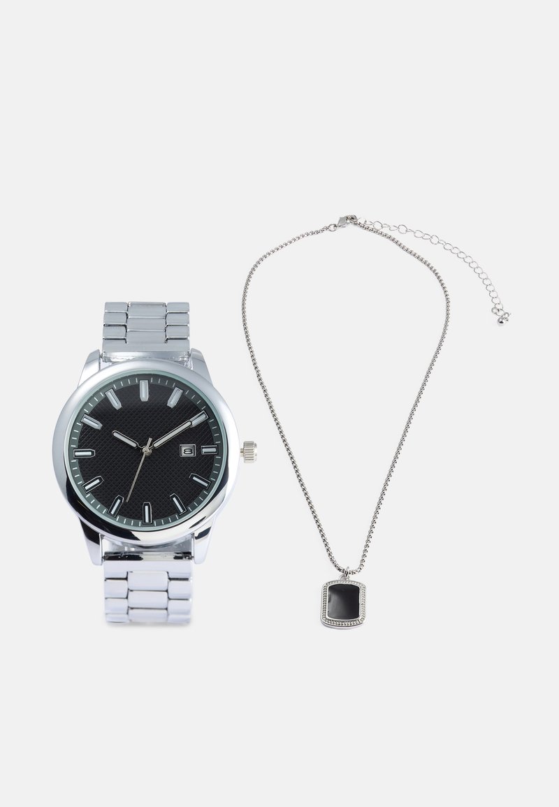 Ezüst óra fekete, texturált számlappal és rozsdamentes acél szíjjal, mellette egy ezüst láncnyaklánc téglalap alakú fekete medállal.