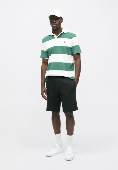 Zaļā un baltā svītraina polo krekls, melni šorti, balti sporta apavi un bēša cepure ar logo. Ikdienišķs apģērbs ar vienkāršām, kontrastējošām krāsām.