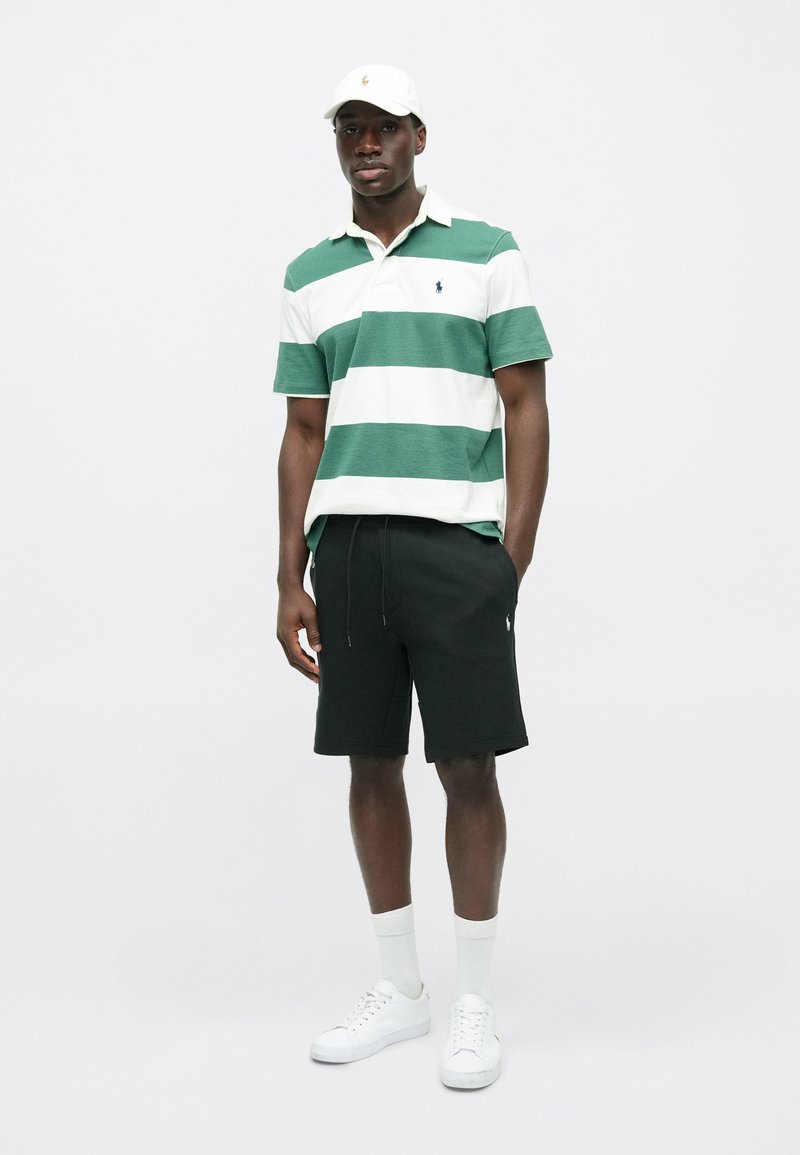 Polo shirt a righe verdi e bianche, pantaloni corti neri, sneakers bianche e un cappellino beige con un logo. Outfit casual con colori semplici e contrastanti.