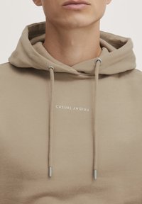 Beige hoodie med snoretræk i hætten. Har en lomme foran og teksten "CASUAL FRIDAY" trykt i hvid. Glat stofstruktur.
