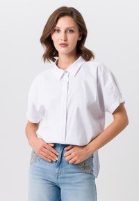 Witte kortarmige blouse met een knoopsluiting aan de voorkant en een puntige kraag, gecombineerd met lichtblauwe jeans met decoratieve kraalwerk op de zakken.