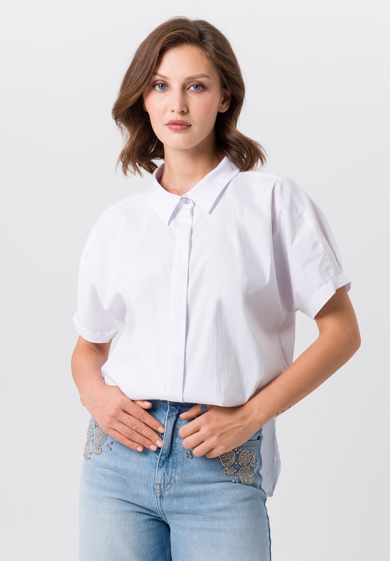 Witte kortarmige blouse met een knoopsluiting aan de voorkant en een puntige kraag, gecombineerd met lichtblauwe jeans met decoratieve kraalwerk op de zakken.
