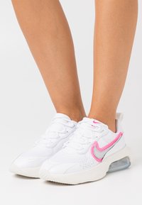White athletic shoes with pink accents, perforated texture, and a visible air cushioning unit. Features a lace-up design and logo detail.