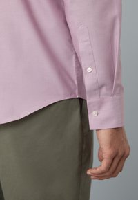 Cămașă roz deschis cu mâneci lungi, manșete cu nasturi, tiv drept și material moale, neted. Purtați cu pantaloni verde măsliniu, evidențiind o croială lejeră.