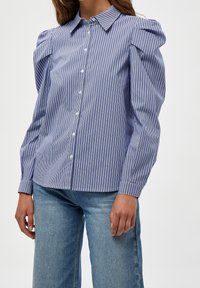 Camisa de rayas azul y blanca con hombros abullonados, botones al frente y un cuello clásico; fabricada con una tela ligera y texturada.