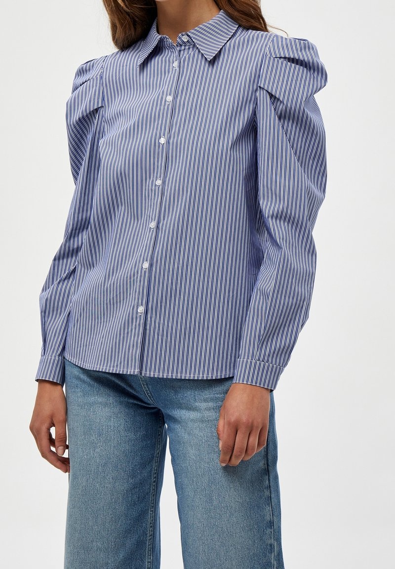 Camisa de rayas azul y blanca con hombros abullonados, botones al frente y un cuello clásico; fabricada con una tela ligera y texturada.