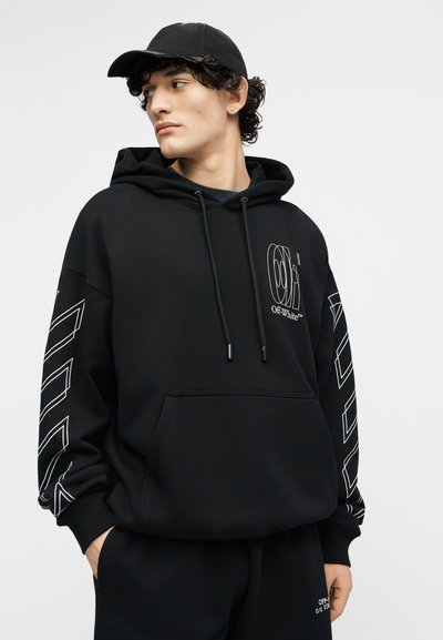 Jauns vīrietis valkā melnu cepuri un melnu kapuci ar baltām ģeometriskām dizainparaugām un uzrakstu "Off-White", skatās uz kreiso pusi ar rokām kabatās.