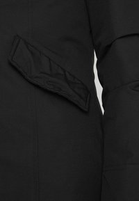 Sort overtøj med et tekstureret stof og et karakteristisk foldet klapdetalje på siden, der fremhæver dets moderne design.