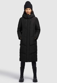 Nuori nainen, joka on pukeutunut pitkään mustaan untuvatakkiin, mustaan neulottuun myssyyn, mustiin legginsseihin ja mustiin nauhavarrellisiin saappaisiin, seisoo eteenpäin käännettynä.