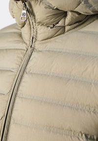 Beige Puffermantel mit horizontalen Steppnähten, hohem Kragen und silbernem Reißverschluss. Die Textur ist glatt mit matter Oberfläche.