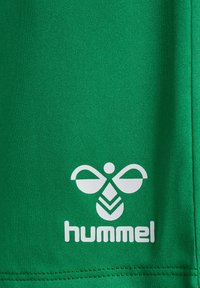 Zielony strój sportowy wykonany z teksturowanej tkaniny, z białym logo o symetrycznym wzorze i nazwą marki "hummel" na dolnej krawędzi.