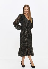 Czarna sukienka w kropki z przezroczystymi długimi rękawami, dekoltem w kształcie litery V oraz falbaniastym wykończeniem na ramionach i u dołu, przewiązana w talii paskiem.