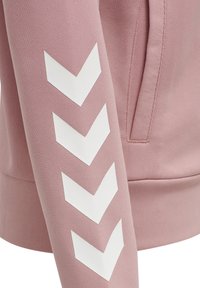 Ärmel eines pinkfarbenen Sweatshirts mit vier großen weißen nach unten zeigenden Chevron-Streifen und einer Seitennähtasche mit sichtbaren Nähten.