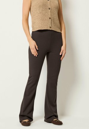 Kvinde iført brun, knappet, ærmeløs striktop, mørkebrune udvidede bukser og matchende mørkebrune flade sko, stående mod en ensfarvet baggrund.