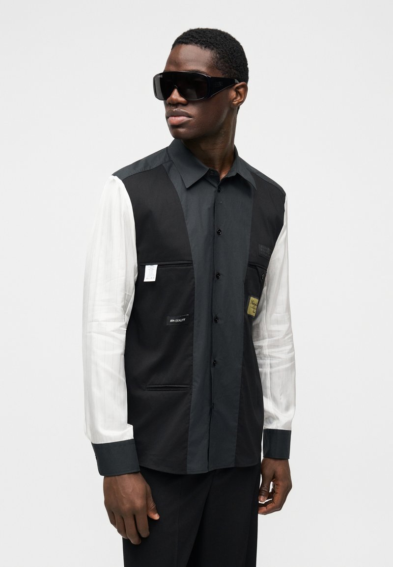 Chemise noir et blanc avec un design à boutons, une manche gauche rayée contrastante, plusieurs poches et une finition texturée.