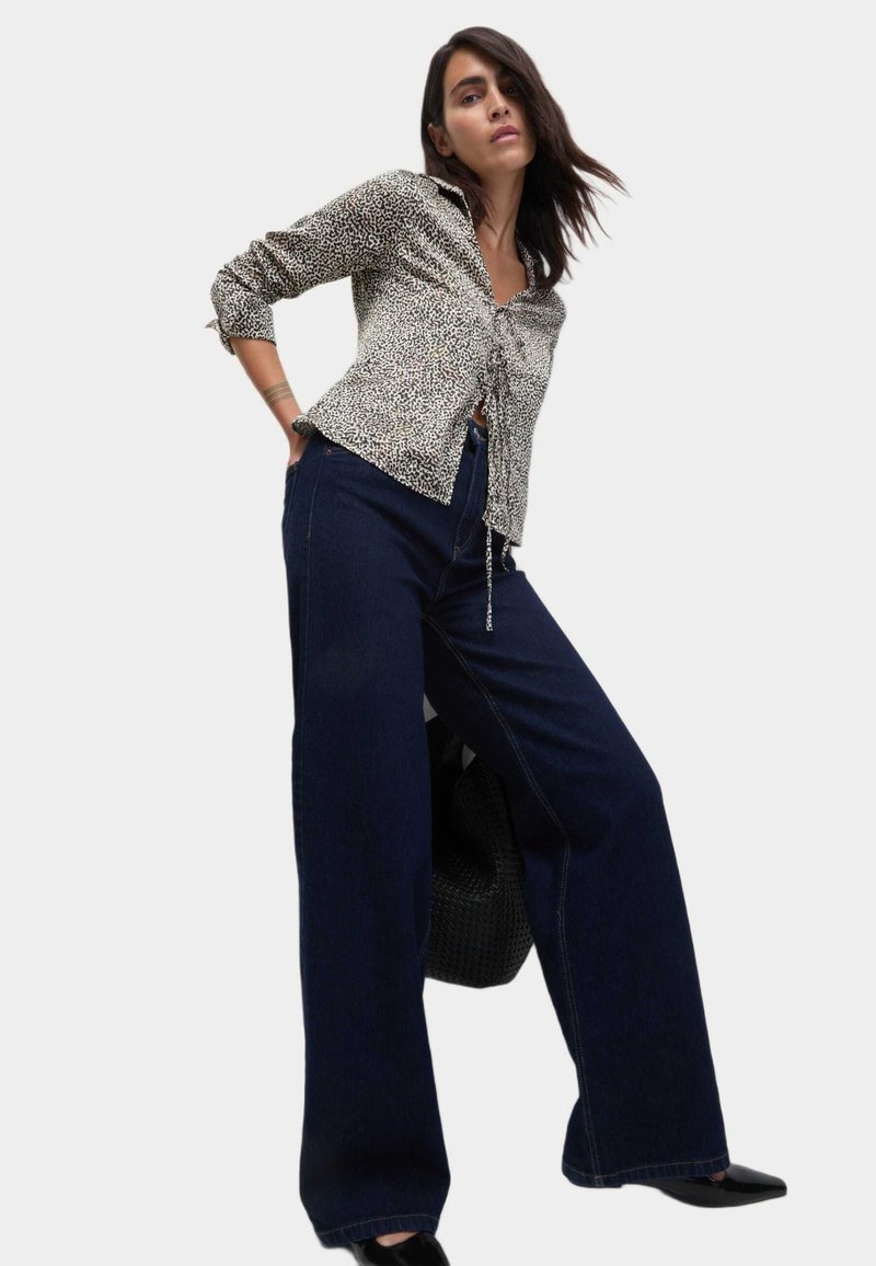 Blusa impresa en blanco y negro con mangas largas y lazo en el frente, combinada con pantalones de mezclilla de pierna ancha de color azul oscuro. La modelo posa con un bolso.