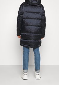 Abrigo acolchado de color azul marino oscuro con secciones horizontales acolchadas, capucha y mangas negras. Combinado con vaqueros azul claro y zapatillas blancas.