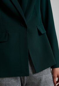 Tmavě zelený dokonale padnoucí blejzr s klopovým límcem, zapínáním na jeden knoflík a klopovými kapsami, v kombinaci se šedými kostkovanými kalhotami.