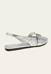 Sandalo slingback metallico argento con accenti borchiati e fibbia. Suola piatta con una texture liscia e un design elegante.