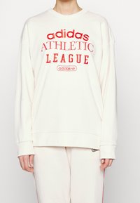 Crème kleurige sweatshirt met geborduurde rode tekst met de woorden "adidas ATHLETIC LEAGUE" en logo. Losse pasvorm met verlaagde schouders.