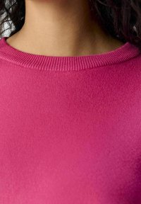 Dopasowany sweter w jasnoróżowym kolorze z żebrowanym okrągłym dekoltem i gładką teksturą. Widok z bliska podkreśla blask tkaniny i intensywność koloru.