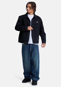 Νέος άντρας με πλεξουδωτά μαλλιά, που φοράει μαύρο τζιν μπουφάν, λευκό πουκάμισο, φαρδιά μπλε τζιν και μαύρα παπούτσια, κοιτάζοντας προς τα πλάγια.