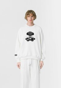 Sudadera blanca con bordado de un oso de peluche en negro y gris en la parte delantera, con puños y dobladillo de canalé, combinada con pantalones blancos.