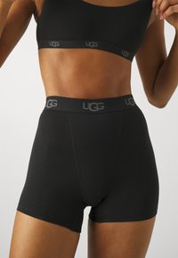 Crne UGG bokserice s glatkom teksturom i elastičnom trakom za struk, na kojoj se nalazi sivi UGG logo. Dizajn je usko prilagođen i dose�že sredinu butina.