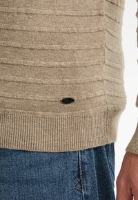 Beige gestrickter Pullover mit strukturierten horizontalen Streifen, geripptem Saum und einem kleinen schwarzen Logo-Tag an der Seite, getragen über blauen Denim-Jeans.