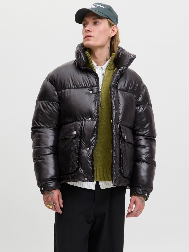 Schwarze Pufferjacke mit glänzender Textur, Druckknöpfen und zwei Fronttaschen. Getragen über einem grünen Pullover und einem weißen Hemd.